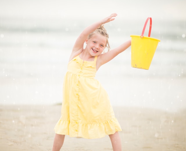 Dziewczynka w żółtej sukience stoi na plaży, trzyma wiaderko